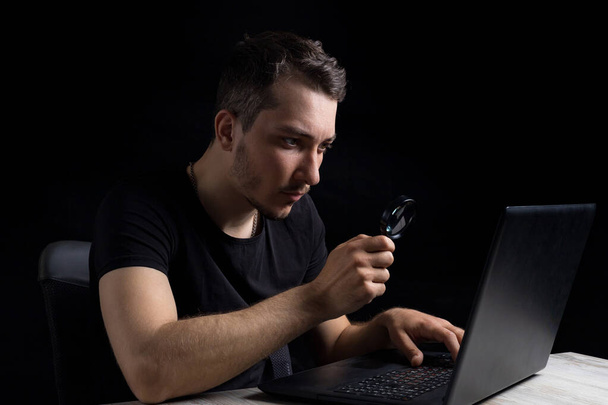 Молодой красивый мужчина смотрит через увеличительное стекло на экран ноутбука на черном фоне в тихом ключе. Поиск символов. Концепция расследования - Фото, изображение