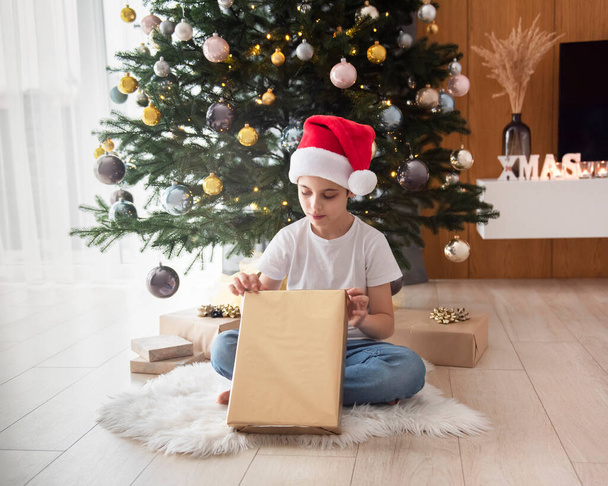 Το αγόρι με τα δώρα παίζει κοντά στο χριστουγεννιάτικο δέντρο. Σαλόνι εσωτερικό με χριστουγεννιάτικο δέντρο και διακοσμήσεις. Πρωτοχρονιά. Δώρο. - Φωτογραφία, εικόνα