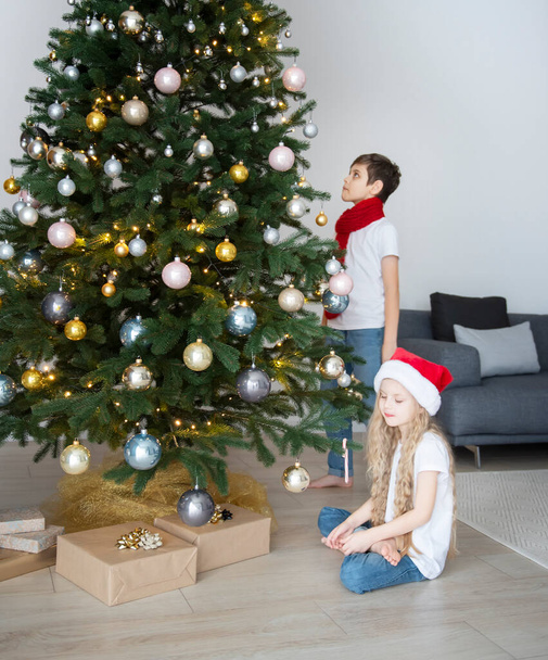 Kinderen - een jongen en een meisje spelen vlakbij de kerstboom. Woonkamer interieur met kerstboom en decoraties. Nieuwjaar. Cadeau geven. - Foto, afbeelding