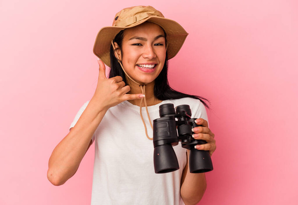 Young mixed race explorer woman holding binoculars isolated on pink background showing a mobile phone call gesture with fingers. - Photo, Image