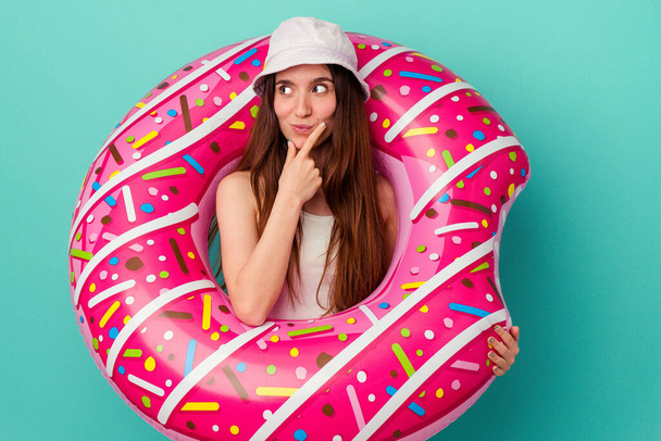 Νεαρή καυκάσια γυναίκα με φουσκωτό ντόνατ απομονωμένη σε μπλε φόντο χαλαρή σκέψη για κάτι κοιτάζοντας ένα χώρο αντιγραφής. - Φωτογραφία, εικόνα