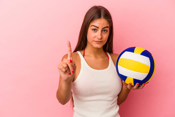 Молода біла жінка тримає волейбольний м'яч ізольовано на рожевому фоні, показуючи номер один пальцем
. - Фото, зображення