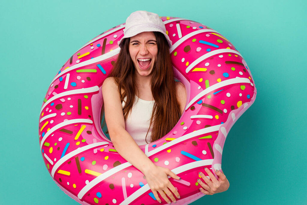 Mulher caucasiana jovem com um donut inflável isolado no fundo azul gritando muito irritado e agressivo. - Foto, Imagem