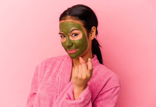 Jeune femme vénézuélienne portant un peignoir et un masque facial isolés sur fond rose pointant du doigt vers vous comme si vous invitant à venir plus près. - Photo, image
