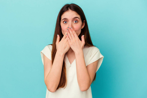 Young caucasian woman isolated on blue background shocked covering mouth with hands. - Фото, изображение