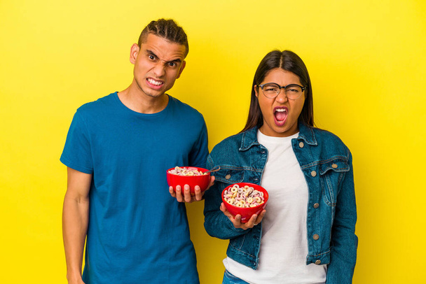 Giovane coppia latina in possesso di una ciotola di cereali isolato su sfondo giallo urlando molto arrabbiato e aggressivo. - Foto, immagini