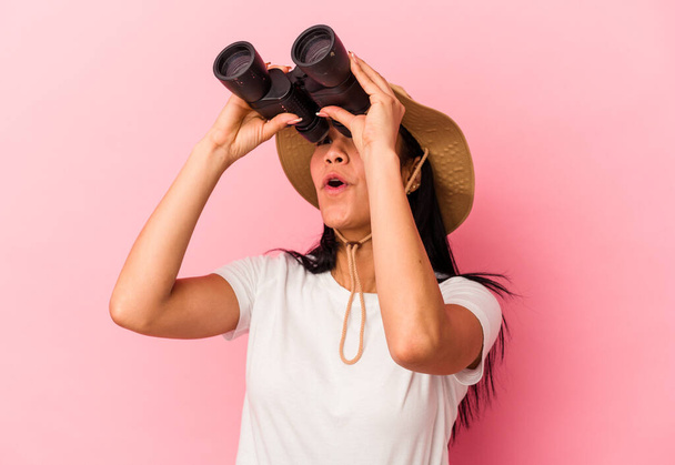 Young mixed race explorer woman holding binoculars isolated on pink background - 写真・画像