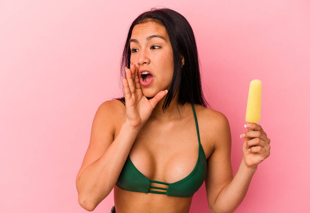 Young Venezuelan woman holding an ice cream isolated on pink background shouting and holding palm near opened mouth. - Foto, Imagen