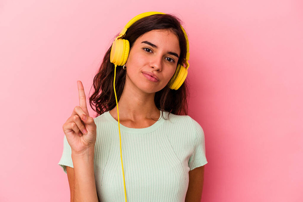 Nuori valkoihoinen nainen kuuntelee musiikkia eristetty vaaleanpunainen tausta osoittaa numero yksi sormella. - Valokuva, kuva