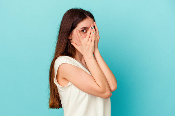 Young caucasian woman isolated on blue background blink through fingers frightened and nervous. - Foto, Imagen