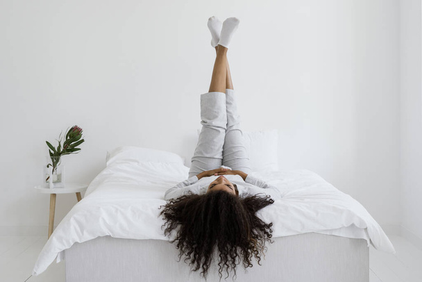 Donna afroamericana felice e distratta sdraiata sul letto in pigiama, con le gambe alzate, che riposava al mattino a casa. Ricreazione, concetto di relax - Foto, immagini