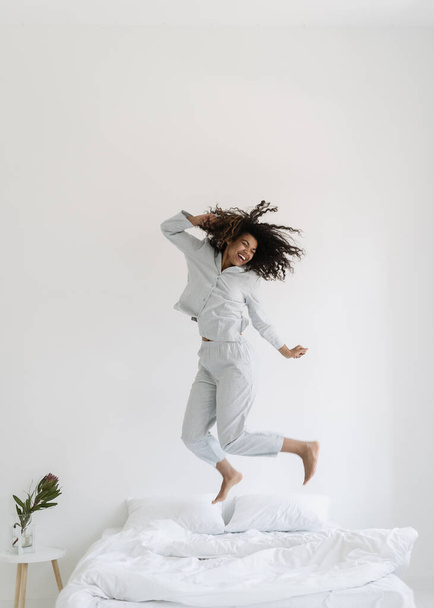 Vertical view of happy mixed race woman dancing in morning, jumping over bed in pajamas. Excited female spending day of in hotel or at home, having fun, feeling joyful mood. Careless, carefree concept - Photo, Image