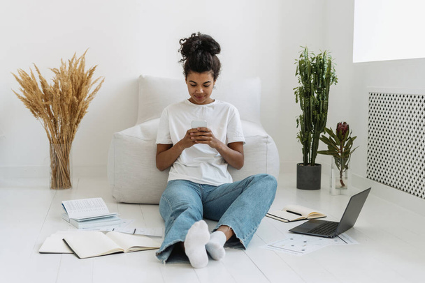 Mladá a klidná studentka v neformálním oblečení sedící na podlaze, používající online aplikaci na svém moderním smartphonu a odpočívající doma po vyučování. Bezdrátová technologie a koncept prohlížení internetu - Fotografie, Obrázek
