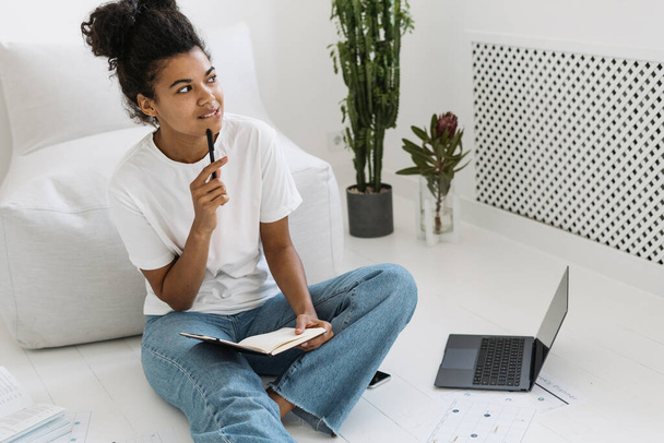 Young and thoughtful millennial girl study at home, writing notes in diary or notepad. Dreamy student girl sitting on floor in bright white room, prepares homework or examination in university - Photo, image