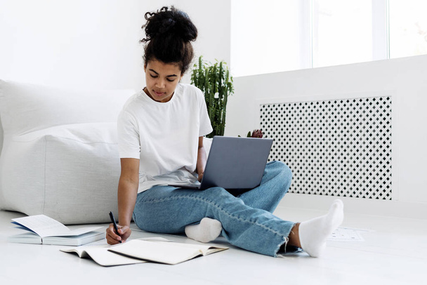 Genç ve zeki kız günlüğe ya da not defterine notlar yazıyor, yerde oturuyor, dizlerinin üzerinde modern dizüstü bilgisayarı tutuyor. Uzaktan çalışma veya serbest çalışma kavramı - Fotoğraf, Görsel