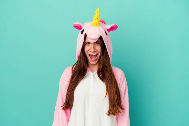 Giovane donna caucasica indossa un pigiama unicorno isolato su sfondo blu urlando molto arrabbiato e aggressivo. - Foto, immagini