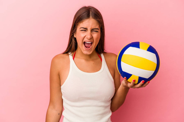 Giovane donna caucasica in possesso di una palla da pallavolo isolato su sfondo rosa urlando molto arrabbiato e aggressivo. - Foto, immagini