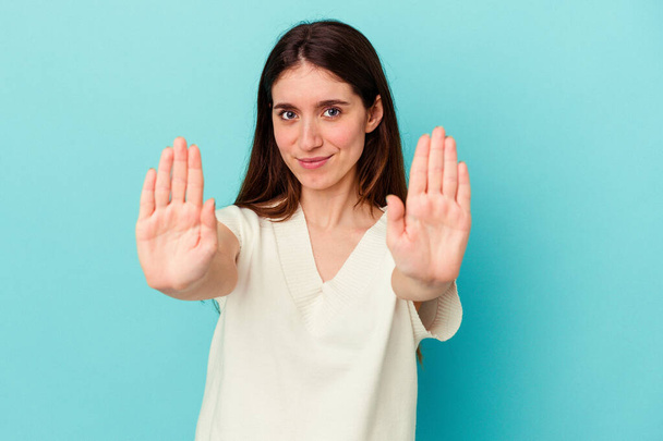 Nuori valkoihoinen nainen sinisellä taustalla seisoo ojennetulla kädellä osoittaa stop-merkki, estää sinua. - Valokuva, kuva