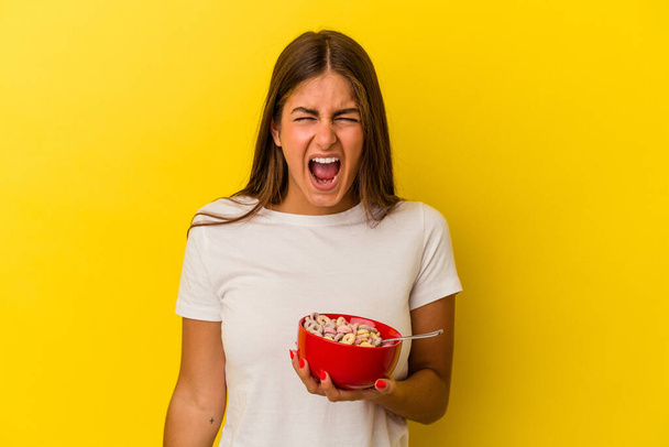 Giovane donna caucasica che tiene i cereali isolati su sfondo giallo urlando molto arrabbiato e aggressivo. - Foto, immagini