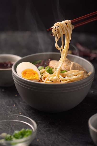 Steam rises over asian noodles in a bowl with meat and vegetables or seafood, often flavored with soy sauce or miso. Dark background - Zdjęcie, obraz