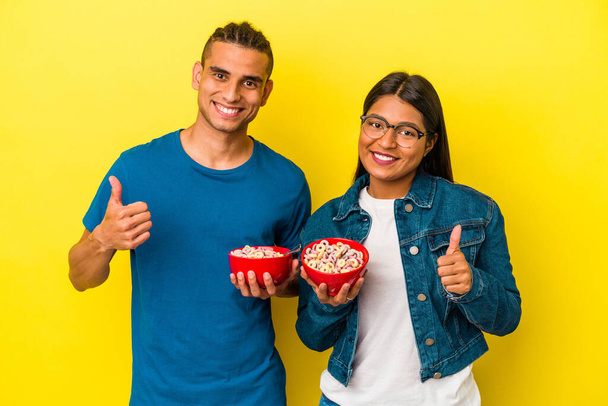 Joven pareja latina sosteniendo un tazón de cereales aislado sobre fondo amarillo sonriendo y levantando el pulgar hacia arriba - Foto, imagen