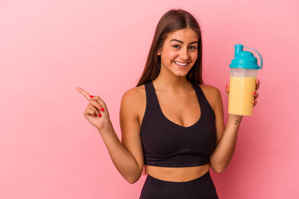 Young caucasian woman holding a protein shake isolated on yellow background smiling and pointing aside, showing something at blank space. - Fotó, kép