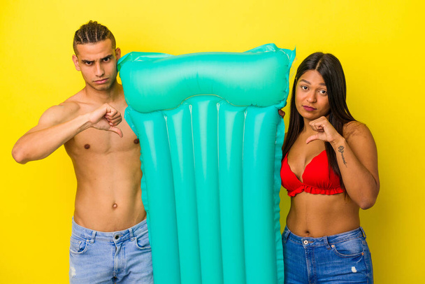 Young latin couple holding air mattress isolated on yellow background showing a dislike gesture, thumbs down. Disagreement concept. - Fotografie, Obrázek