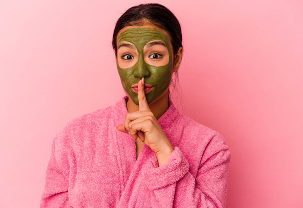 Jeune femme vénézuélienne portant un peignoir et un masque facial isolés sur fond rose gardant un secret ou demandant le silence. - Photo, image