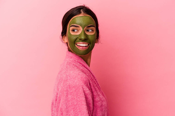 Mujer caucásica joven con una bata de baño y mascarilla facial aislada sobre fondo rosa se ve a un lado sonriente, alegre y agradable. - Foto, imagen