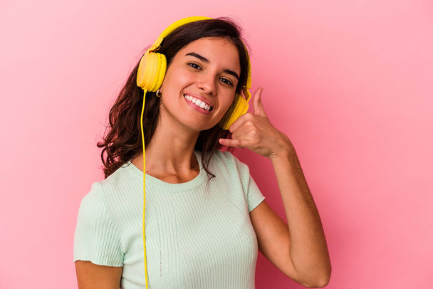 Young caucasian woman listening music isolated on pink background showing a mobile phone call gesture with fingers. - 写真・画像