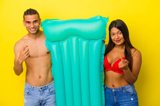 Jovem casal latino segurando colchão de ar isolado no fundo amarelo apontando com o dedo para você como se convidando chegar mais perto. - Foto, Imagem