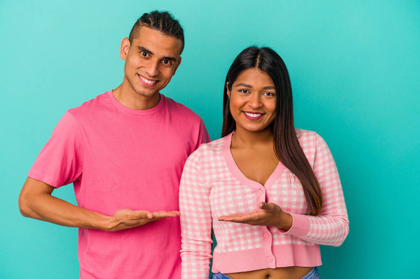 Jovem casal latino isolado no fundo azul mostrando um espaço de cópia em uma palma e segurando outra mão na cintura. - Foto, Imagem