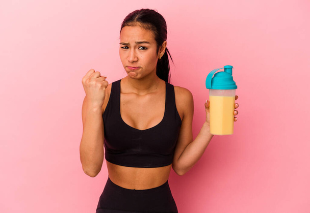 Young Venezuelan woman drinking a protein shake isolated on pink background showing fist to camera, aggressive facial expression. - Photo, Image