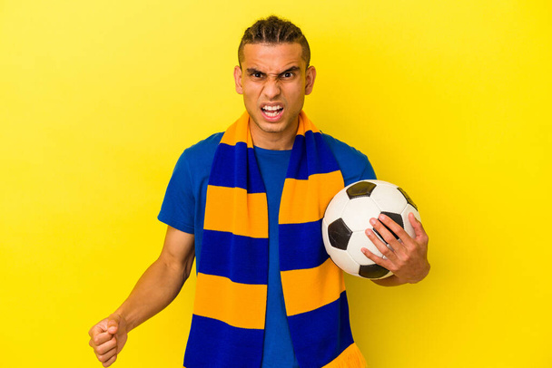 Nuori venezuelalainen mies katsomassa jalkapalloa eristettynä keltaisella taustalla huutaen hyvin vihainen ja aggressiivinen. - Valokuva, kuva