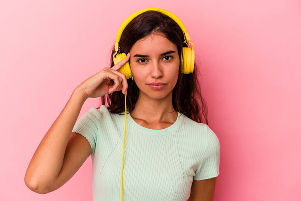 Nuori valkoihoinen nainen kuuntelemassa musiikkia eristetty vaaleanpunainen tausta osoittaa temppeliä sormella, ajattelu, keskittynyt tehtävään. - Valokuva, kuva