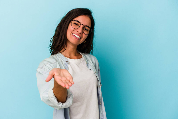 Young caucasian woman isolated on blue background stretching hand at camera in greeting gesture. - Photo, Image