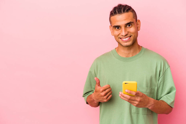 Young Venezuelan man holding a mobile phone isolated on pink background smiling and raising thumb up - Foto, Imagem