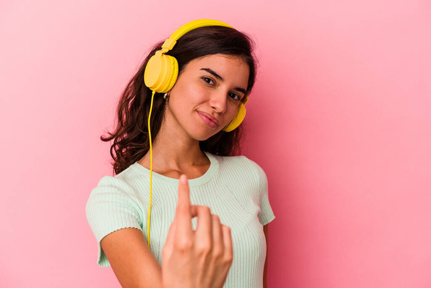 Young caucasian woman listening music isolated on pink background pointing with finger at you as if inviting come closer. - Foto, Imagem