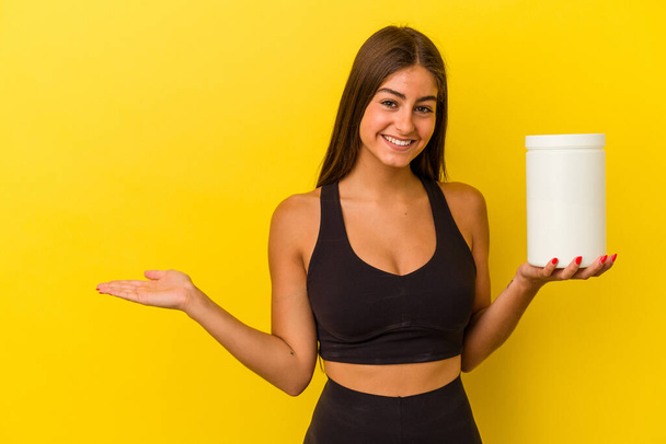 Young caucasian woman holding a protein bottle isolated on yellow background showing a copy space on a palm and holding another hand on waist. - Fotó, kép