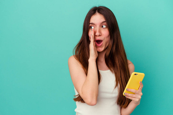 Giovane donna caucasica in possesso di un telefono cellulare isolato su sfondo blu sta dicendo una notizia segreta frenante caldo e guardando da parte - Foto, immagini