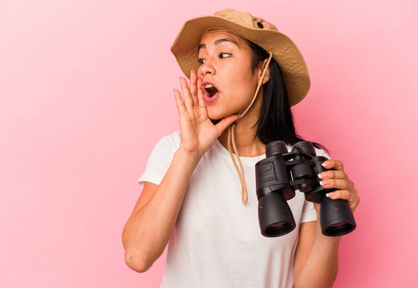 Mladý smíšený závodní průzkumník žena drží dalekohled izolované na růžovém pozadí říká tajné horké brzdící zprávy a dívá se stranou - Fotografie, Obrázek