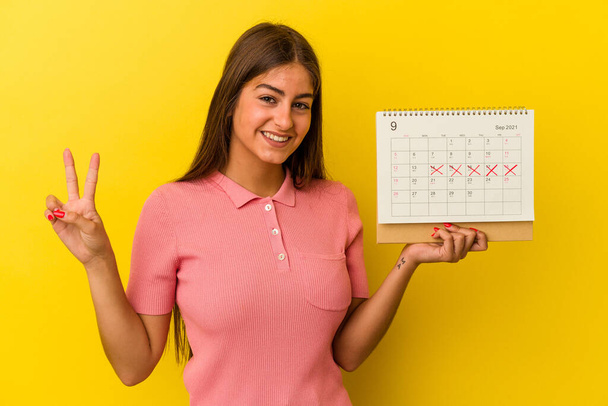 Jeune femme caucasienne tenant un calendrier isolé sur fond jaune montrant le numéro deux avec les doigts. - Photo, image