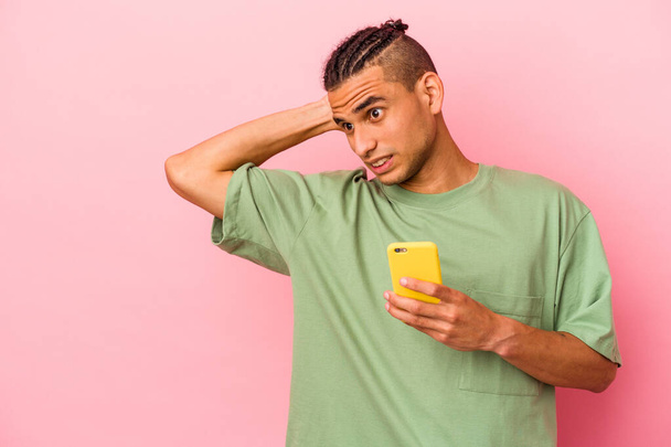 Молодий венесуельський чоловік, що тримає мобільний телефон ізольований на рожевому фоні, торкаючись потилиці, думає і робить вибір.. - Фото, зображення