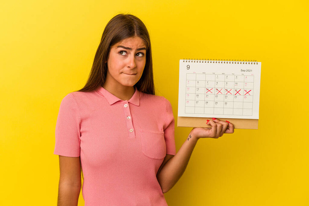 黄色の背景に孤立カレンダーを保持若い白人女性が混乱し、疑問と不確定な感じ. - 写真・画像