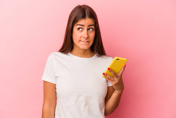 Junge kaukasische Frau hält ein Handy isoliert auf rosa Hintergrund verwirrt, fühlt sich zweifelhaft und unsicher. - Foto, Bild