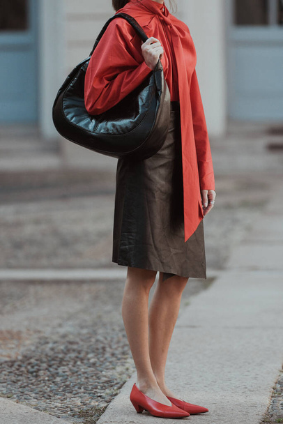 Milan, Italy - September 24, 2021:  Street style outfit, fashionable woman wearing  a red oversized wrap shirt with a knotted neck, a black shiny leather shoulder bag, a black shiny leather high waist knees skirt on the streets of Milan, Italy. - Zdjęcie, obraz