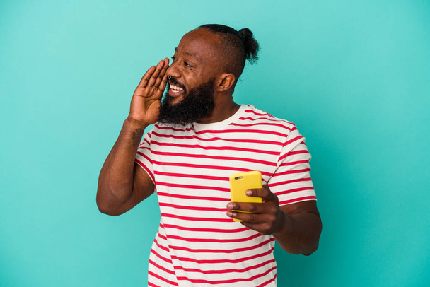 Afrykański Amerykanin trzyma telefon komórkowy odizolowany na niebieskim tle krzycząc i trzymając dłoń blisko otwartych ust. - Zdjęcie, obraz