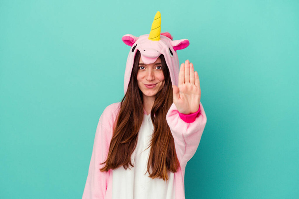 Mujer caucásica joven con un pijama de unicornio aislado sobre fondo azul de pie con la mano extendida mostrando señal de stop, lo que le impide. - Foto, imagen