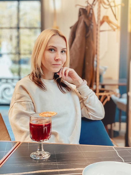 A blonde caucasian joyful girl in a light sweater sits in a restaurant near the window at a table with a mulled wine. - Фото, изображение