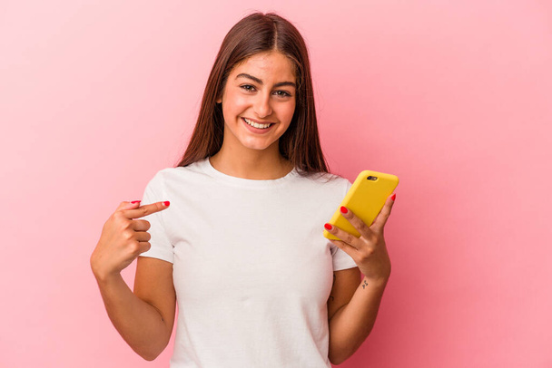 Молода біла жінка тримає мобільний телефон ізольовано на рожевому фоні, вказуючи вручну на простір для копіювання сорочки, пишаючись і впевнено
 - Фото, зображення